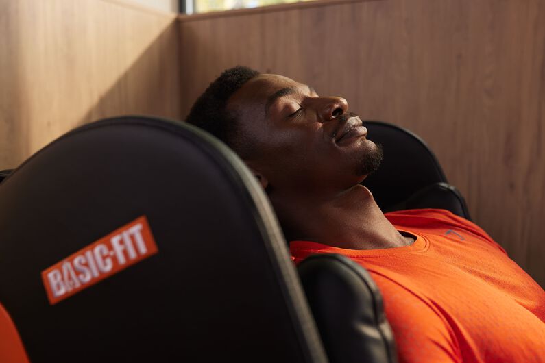 Een man in een oranje sportshirt ontspant met gesloten ogen in een zwarte massagestoel met het Basic-Fit logo.