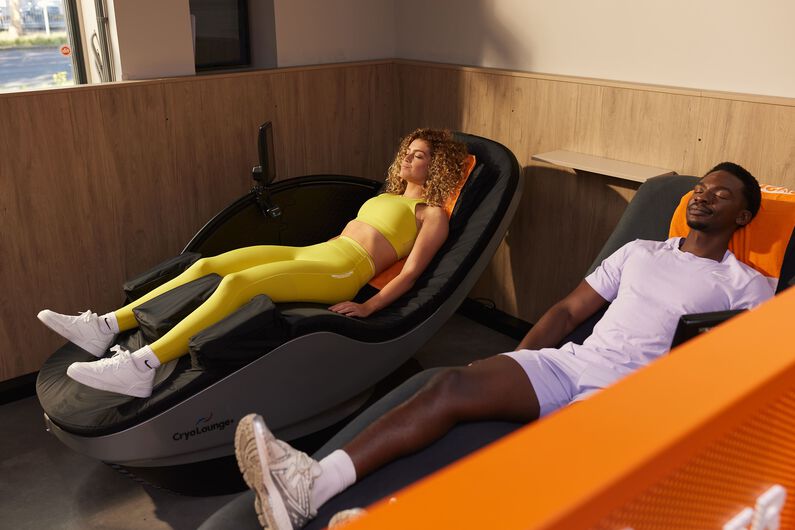 Een vrouw in een gele sportoutfit ontspant op een cryobed, terwijl een man in een lichtpaarse outfit uitrust op een hydromassagebed bij Basic-Fit.