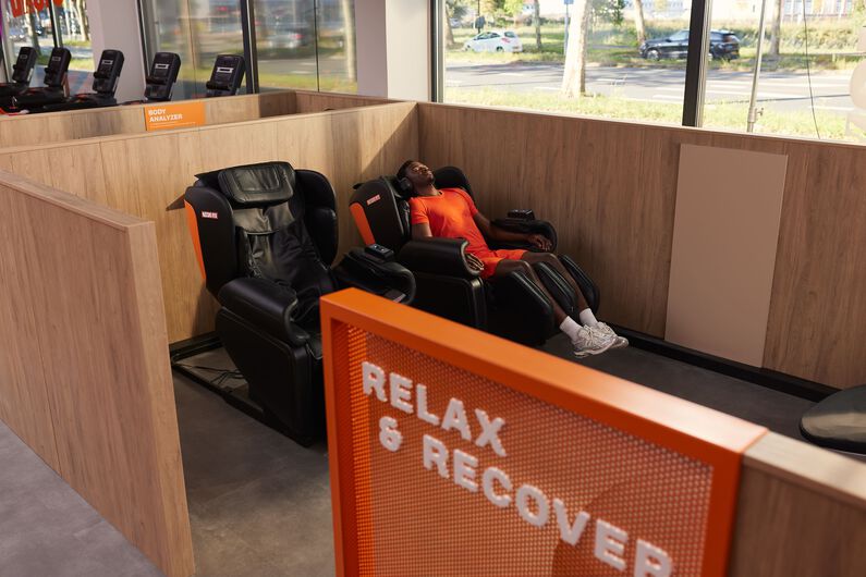 Een man in een rode sportoutfit ontspant met een koptelefoon in een massagestoel in de Relax & Recover zone bij Basic-Fit.