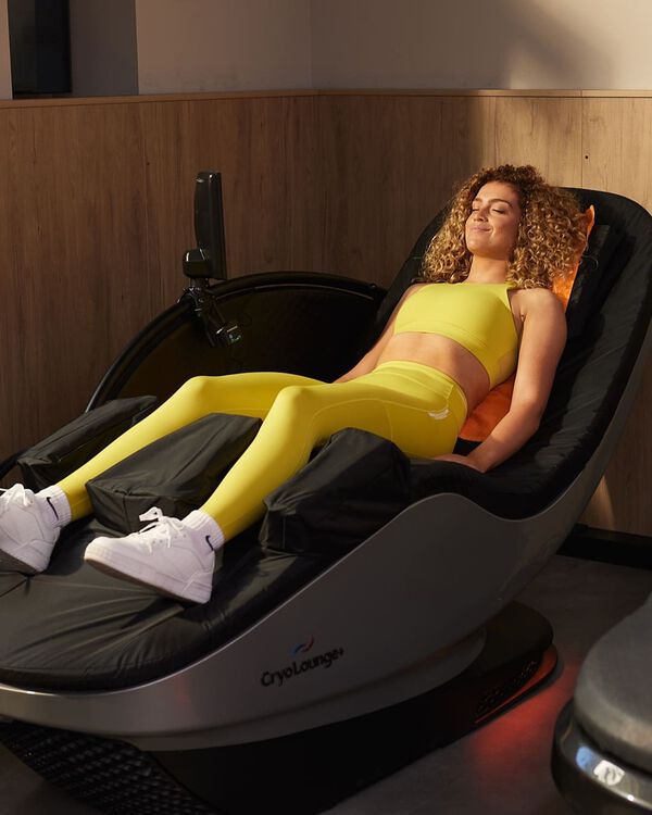 Een vrouw in een gele sportoutfit ontspant op een CryoLounge+ herstelstoel met een oranje handdoek achter haar hoofd bij Basic-Fit.