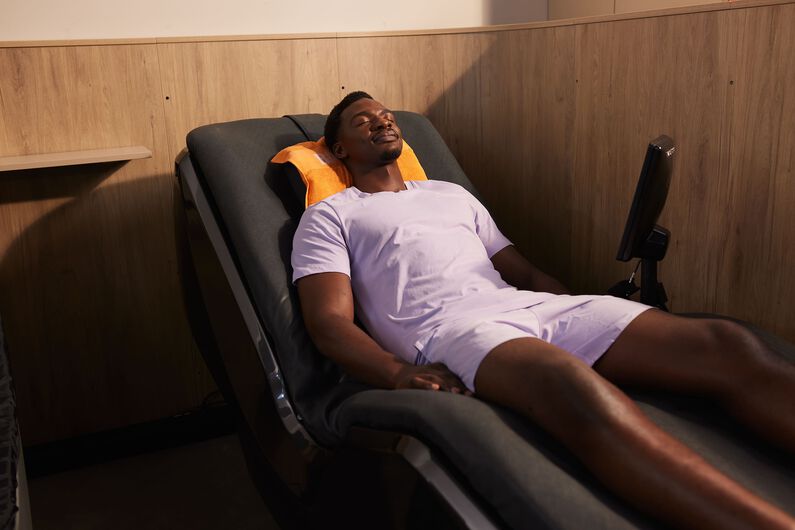 Een man in een lichtpaarse sportoutfit ontspant op een hydromassagebed met een oranje handdoek achter zijn hoofd bij Basic-Fit.