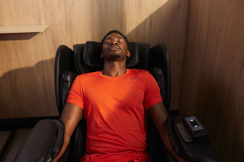 Een man in een rode sportoutfit ontspant met gesloten ogen in een massagestoel bij Basic-Fit.